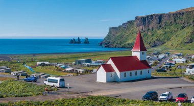 İzlanda, Vik i Myrdal 'da kırmızı çatılı beyaz bir kilisenin hava manzarası. Kilise arka planda siyah kumsal, okyanus ve yeşil tepelere karşı duruyor..
