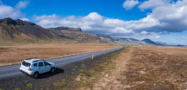 Beyaz bir SUV, İzlanda 'da beyaz bulutlu mavi bir gökyüzünün altında kuru otlar ve küçük tepelerle çevrili asfalt bir yolda ilerliyor. Manzaralı güzergahın hava görüntüsü.