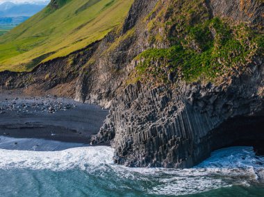 Dramatic aerial image of a black sand beach in Iceland, bordered by cliffs with unique basalt columns. Waves crash against the shore, creating dynamic energy. clipart