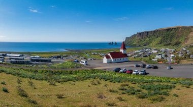 Vik i Myrdal, İzlanda 'nın ikonik siyah kumsal sahili, kırmızı çatılı beyaz kilise ve uzak mesafedeki Reynisdrangar deniz yığınları..