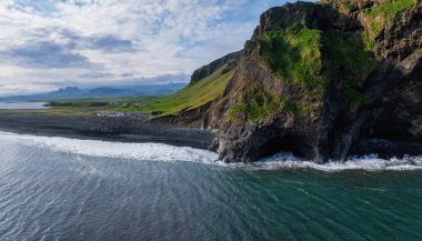 İzlanda kıyı şeridinin çarpıcı hava manzarası bir mağara, yemyeşil bitki örtüsü, siyah bir kumsal ve turkuaz okyanus suları ile dramatik bir uçurum sergiliyor..