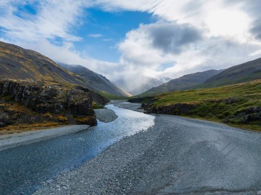 Geniş, sığ bir nehir, çakıl yataklı, yemyeşil bir vadi boyunca esiyor. Etrafı kar kaplı dağlarla çevrili. Mavi gökyüzünün altında beyaz bulutlar var..
