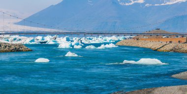 İzlanda 'da karla kaplı dağlarla çevrili yüzen buz kütleleriyle dolu bir nehri kaplayan asma köprünün hava görüntüsü. Buzlu sularla kaplı kış manzarası.