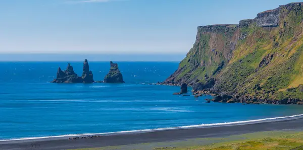 stock image Stunning aerial view of a black sand beach in Iceland, bordered by rocky cliffs and distant sea stacks. The vibrant blue water and clear sky enhance the dramatic landscape.