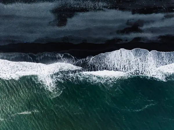 stock image Dramatic aerial view of Icelands black sand beach with striking contrast between dark sand and white foamy waves crashing against the shore.