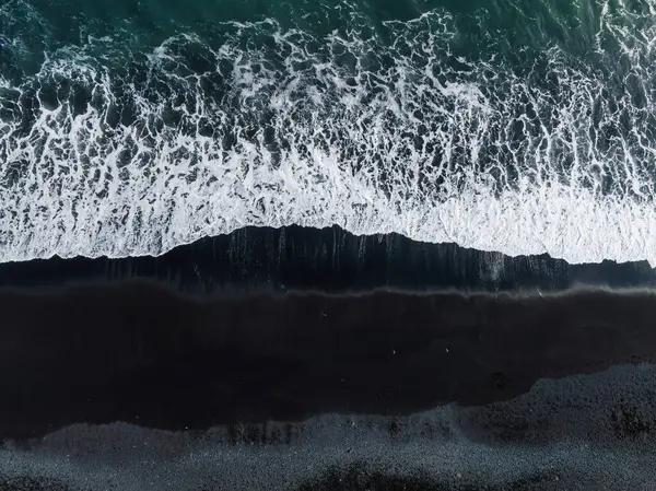 stock image Dramatic contrast of white ocean waves crashing against the black sand beach in Iceland. Captured from above, showcasing the unique natural beauty.