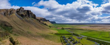 İzlanda 'nın göz kamaştırıcı hava manzarası. Dik kayalık bir kayalık ve geniş yeşil ovalar. Parlak mavi bir gökyüzünün altında dolambaçlı bir yol ve tüylü beyaz bulutlar..