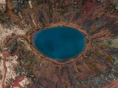 İzlanda 'daki volkanik bir krater gölü, koyu mavi sular, kırmızımsı kahverengi bir çember ve çevresindeki çeşitli arazilerin sersemletici hava görüntüsü..