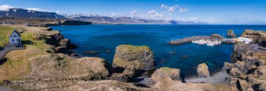 İzlanda 'nın engebeli kıyı şeridi, göz kamaştırıcı hava manzarası. Mavi gökyüzü altında küçük beyaz bir ev, derin mavi sular ve uzak bir liman..