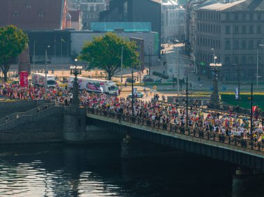 Riga, Letonya 'daki bir maratonun enerjisini yansıtan canlı bir hava çekimi. Koşucular, binalar ve yemyeşil ağaçlarla çevrili bir nehri geçerek köprüden geçiyorlar..