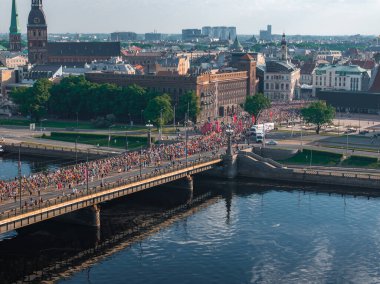 Riga, Letonya 'da bir nehrin üzerinden geçen büyük bir kalabalığın enerji dolu görüntüsü. Şehir silüeti maraton etkinliği için çarpıcı bir zemin oluşturuyor..