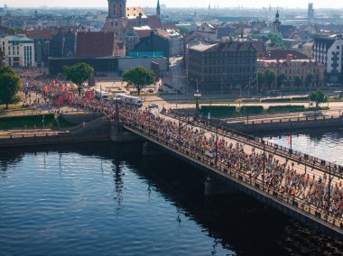 Canlı bir hava görüntüsü, Riga, Letonya 'da bir köprüden geçen büyük bir maraton koşucusunu yakalıyor. Arka planda şehrin ufuk çizgisi var..