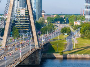 Riga, Letonya 'da kablo destekli bir köprüde maraton koşan büyük bir kalabalığın hava görüntüsü. Seyirciler, arka planda şehir manzarası olan köprüyü çiziyor.