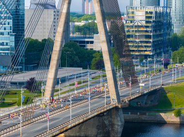 Riga, Letonya 'da kablo destekli bir köprüden geçen maraton koşucularını gösteren dinamik bir çekim. Yüksek binalarla şehir silueti çarpıcı bir zemin oluşturuyor..