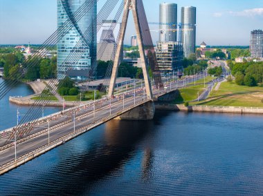Riga, Letonya 'da kablo destekli köprüden geçen büyük bir kalabalığın hava görüntüsü. Şehrin ufuk çizgisi ve durgun nehir mavi gökyüzünü yansıtıyor..