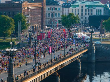 Koşucular Letonya 'nın Riga kentindeki sakin bir nehirden karşıya geçerken, kenarlar seyircilerle çevrili. Arkaplanda eski ve yeni mimarinin bir karışımı yer alıyor.
