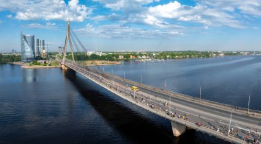 Riga, Letonya 'da kablo destekli bir köprüden geçen maraton koşucularını canlı bir şekilde çeken bir çekim. Durgun nehir ve mavi gökyüzü beyaz bulutlarla sahnelere enerji katar.