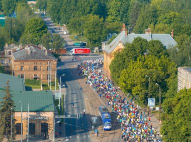 Riga Rimi Maratonu 2024 'e katılan çeşitli koşucuların enerji dolu sahnesi. Seyirciler sokakları dolduruyor ve Riga şehrinin silueti arka planda görünüyor..