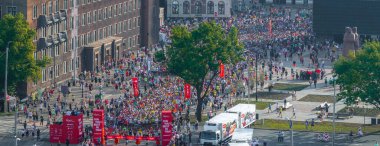 Riga, Letonya 'daki bir maratonun enerjisini yansıtan canlı bir hava çekimi. Renkli kıyafetler giymiş koşucular ve seyirciler sokaklarda coşkuyla tezahürat yapıyorlar..