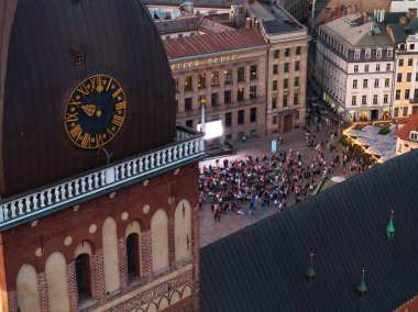 Riga Katedrali 'nin saat kulesinin kuş bakışı görüntüsü ve aşağıdaki meydanda toplanan kalabalık mimari detayları ve hareketli aktiviteleri vurguluyor..