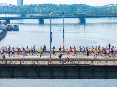 Letonya, Riga 'da açık mavi bir nehrin üzerinde tahta bir güverte ile çelik bir köprüyü geçen büyük bir grup koşucunun hava görüntüsü..