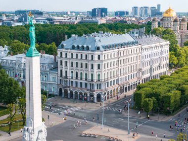 Riga, Letonya 'daki Özgürlük Anıtı' nın kuş bakışı görüntüsü. Birkaç kişi boş sokakta yürüyor ve koşuyor. Arka planda büyük beyaz bir cephe binası görünür..
