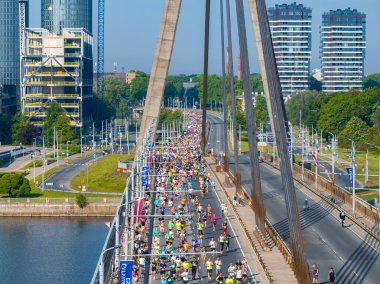 Riga, Letonya 'da bir maratonun havadan çekilmiş görüntüleri. Binalar ve ağaçlarla çevrili, merkezi beton direkli, kablo destekli bir köprüden geçen koşucular görülüyor..