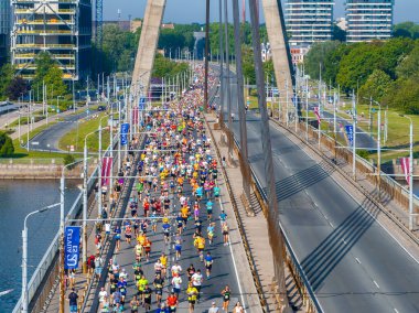Riga, Letonya 'daki bir maratonun havadan çekimi. Çelik bir köprüde koşuculardan oluşan büyük bir kalabalık var. Köprü binalar ve ağaçlarla çevrili. Koşucular renkli giyinmiş..