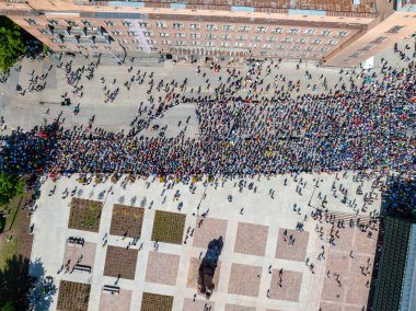 Riga 'daki bir şehir meydanında kalabalık bir kalabalığı çeken hava görüntüleri. İnsanlar parlak renkli kıyafetler giyiyorlar, arka planda binalar ve ağaçlar var..