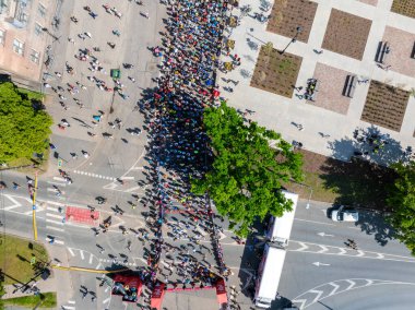 Riga Rimi Maratonu 2024 'e katılan çeşitli koşucuların enerji dolu sahnesi. Seyirciler Riga, Letonya sokaklarında kuyruk oluştururken, arka planda şehrin silüeti görülüyor..