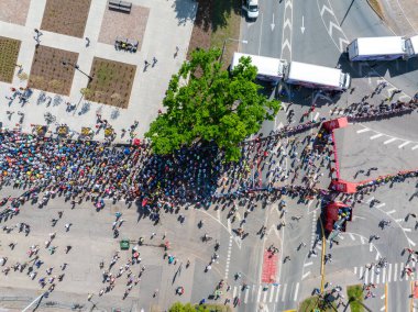 Riga Rimi Maratonu sırasında Riga 'da bir şehir caddesini büyük bir koşucu kalabalığı dolduruyor. Sokak ağaçlar, binalar ve park edilmiş araçlarla dolu..