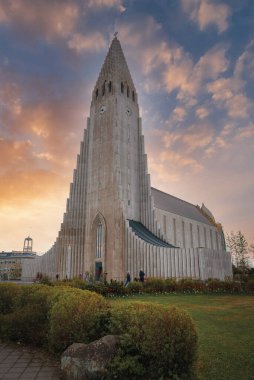 Hallgrimskirkjas benzersiz mimari bazalt sütunlardan esinlenilmiştir, gün batımında sıcak renklerle yakalanmıştır. Sahnede bahçeler ve girişe yakın insanlar var..