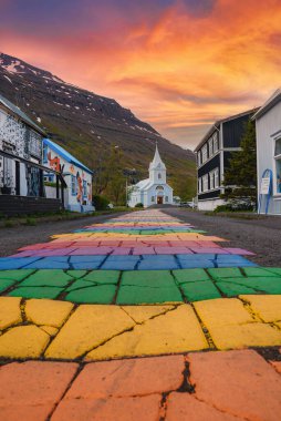 Seydisfjordur, İzlanda 'da canlı bir sahne, gökkuşağı boyalı bir patika ile beyaz bir kiliseye giden, gün batımının alevli arkaplanı altında bir dağ manzarası..