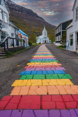 Picturesque street in Seydisfjordur, Iceland, with a rainbow-colored path to a white church. Charming buildings and a mountainous backdrop under a dramatic sky. clipart