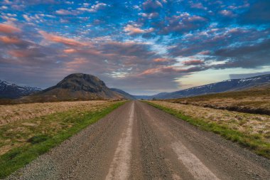 Uzağa uzanan çakıllı bir yol, çimenli tarlalarla çevrili ve İzlanda 'da canlı, parçalı bulutlu bir gökyüzünün altında öne çıkan, yuvarlak bir dağa doğru uzanıyor..