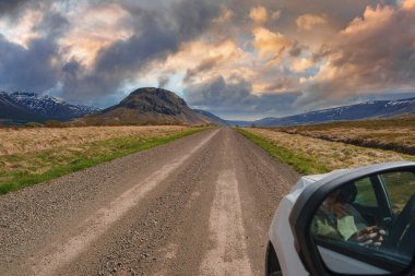 Çimenli tarlaların kuşattığı ve renkli bir gökyüzünün altındaki engebeli bir dağa doğru uzanan çakıllı bir yol uzanıyor. Arka planda karla kaplı tepeler görülebilir.