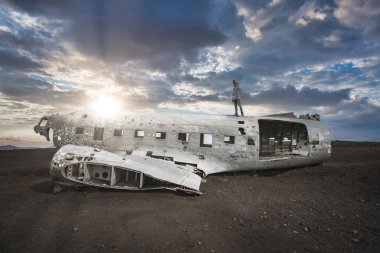 İzlanda, Solheimasandur Sahili 'ndeki Amerikan Donanması Douglas DC-3 enkazının dramatik bir sahnesi. Bulutlu bir gökyüzünün altında güneş ışığı altında bir insan uçağın gövdesinde duruyor..
