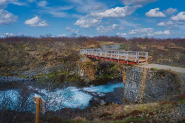 İzlanda 'da bir yürüyüş yolunda küçük ahşap bir köprünün, engebeli arazinin ortasında berrak bir mavi nehri ve parlak bir gökyüzünün altında seyrek bir bitki örtüsünün manzarası..