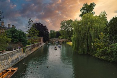 Cambridge, İngiltere 'deki River Cam' in sakin bir sahnesinde, insanların zıpladığı tarihi bir taş köprü, yemyeşil bir köprü ve dramatik bulutlarla sıcak bir günbatımı gökyüzü..