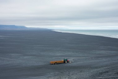 İzlanda 'da geniş siyah bir kumsalda yalnız bir traktör ve karavan. Kumsal, kıyı şeridi boyunca uzanan uzak dağlarla, bulutlu bir gökyüzünün altında okyanusla buluşuyor..