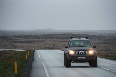 Islak, çift şeritli bir yolda, İzlanda 'da sarı işaretlerle çevrili tek bir SUV bulutlu bir gökyüzü altında ıssız, kayalık bir arazide yol alır..