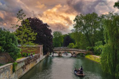 Cambridge, İngiltere 'deki River Cam' in sakin bir sahnesinde muhteşem bir taş köprü, yemyeşil bir köprü ve dramatik bir günbatımı gökyüzü altında uçan bir tekne yer alıyor..