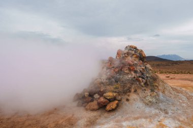 Bir jeotermal buhar bacası İzlanda 'da çorak, kırmızımsı-kahverengi bir alana yoğun buhar yayar. Karla kaplı dağlar ve bulutlu bir gökyüzü benzersiz güzelliği artırır..