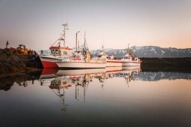 İzlanda 'da balıkçı teknelerinin kenetlendiği ve yansımalarının sakin sularda yansıdığı sakin bir liman. Karla kaplı dağlar ve yumuşak ışık huzurlu ortamı güçlendirir.