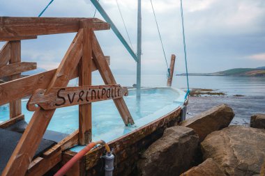 A serene outdoor spa setup featuring a wooden hot tub labeled Sverrins Pollur by a rocky shoreline, with calm waters and distant hills in Iceland. clipart