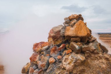 İzlanda 'da bulutlu bir gökyüzünün altında ıssız bir arazide geçen, kırmızımsı kahverengiden gri kayalardan oluşan dumanlı bir fümerol jeotermal aktivite sergiliyor..