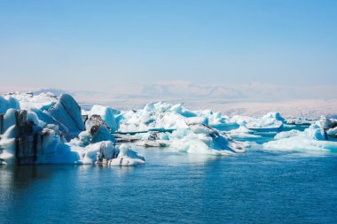 İzlanda 'daki Jokulsarlon Buzul Gölü' nün sakin manzarası, mavi ve beyaz buzdağları, sakin yansıtıcı sular ve berrak bir gökyüzü altında karla kaplı dağlar..