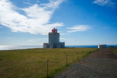 İzlanda 'nın güney kıyısındaki Dyrholaey Deniz Feneri' nin huzurlu manzarası. Kırmızı kubbeli beyaz yapı, çakıl yolu ve dağınık bulutlu açık mavi gökyüzü..