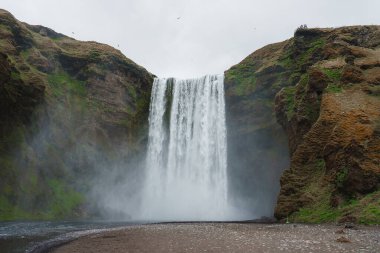 Skogafoss şelalesi İzlanda 'da yosunlu bir uçurumdan aşağı düşer ve sis gizemli bir atmosfer yaratır. Kuşlar tepeye yakın uçar ve platformdaki insanlar onun ölçeğini vurgular..