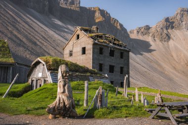 Yıkılan çatısı, Viking heykeli ve İzlanda 'da kayalık dağları olan yıpranmış bir bina. Ahşap yapıları ve piknik masası olan, İzlanda mirasının sergilendiği otlak bir alan..